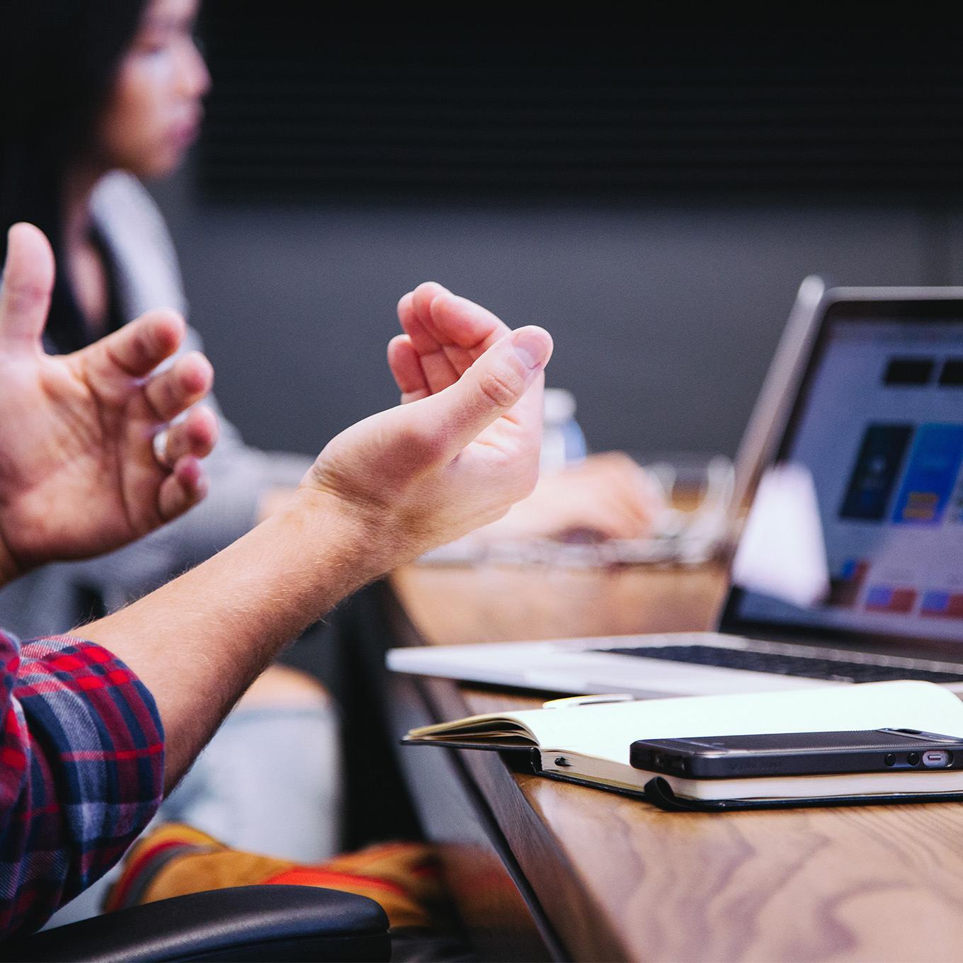 Close up of hands gesticulating, in front of a laptop, pen and notebook.