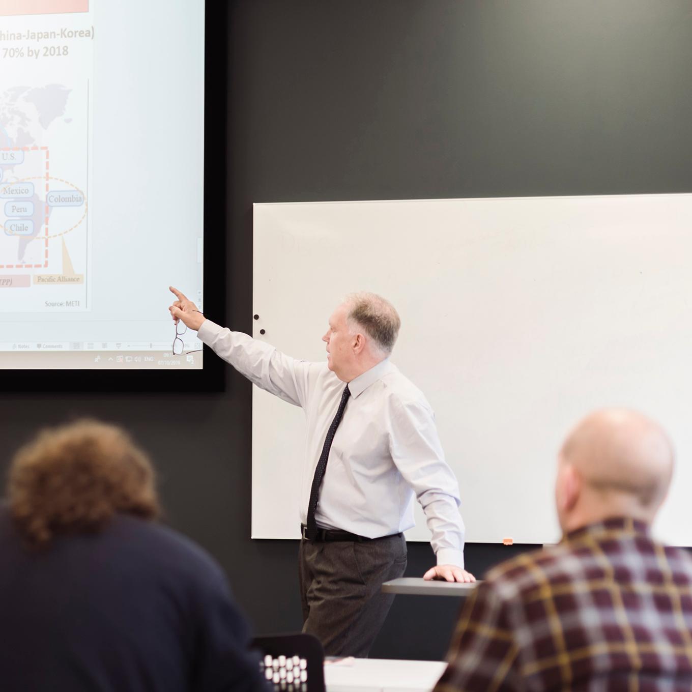 Students in a lecture - the lecturer is pointing at the projection of the presentation slides, while students take notes