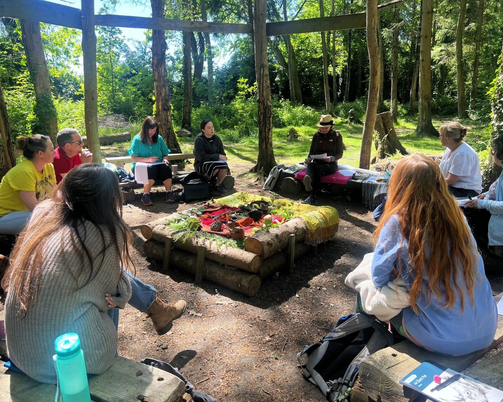 A photo of a group of adults sitting in the forest in a circle, some with sketchpads.