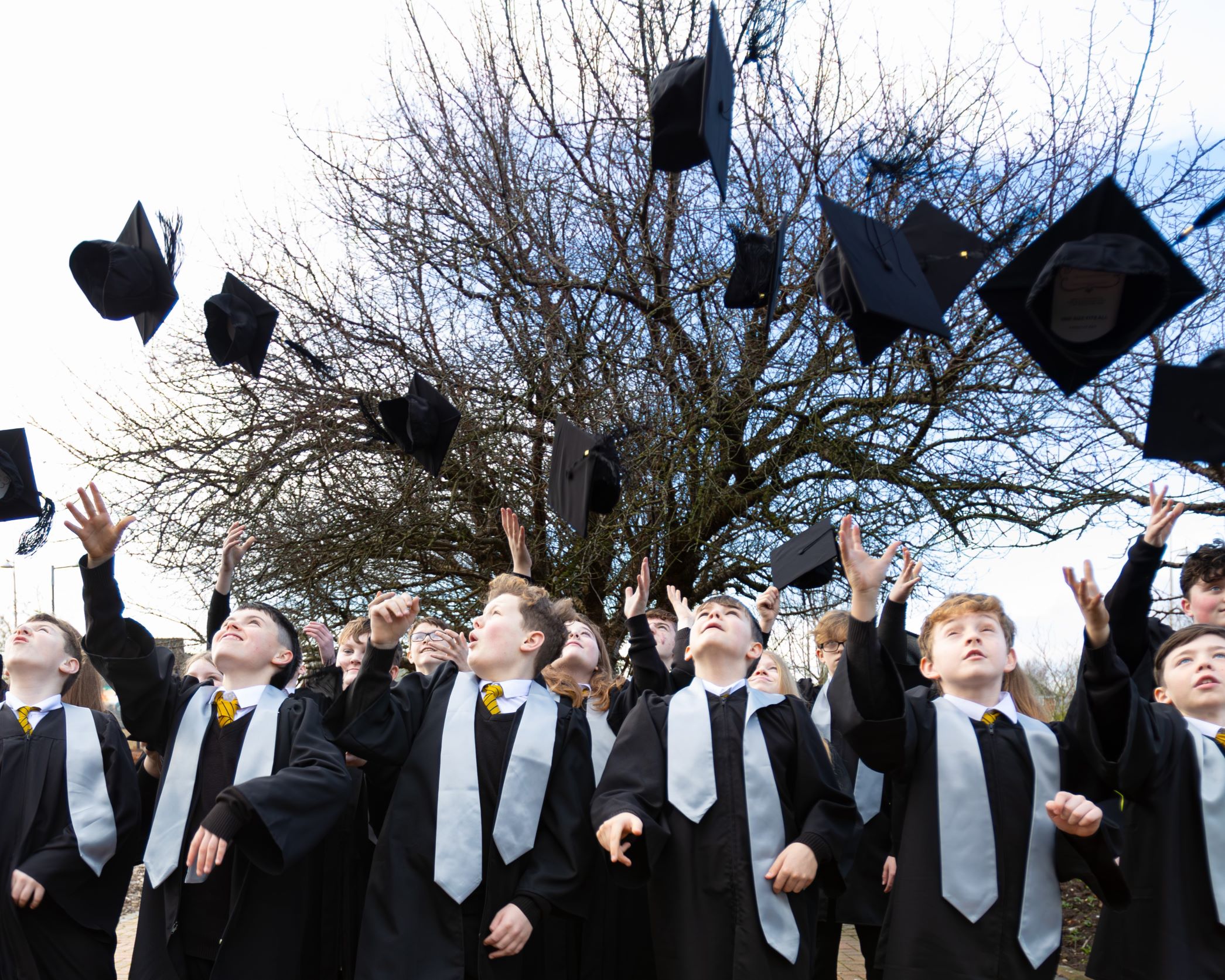 A group of young people graduating the Children's University programme in a wearing a graduation gown and throwing their caps into the air.