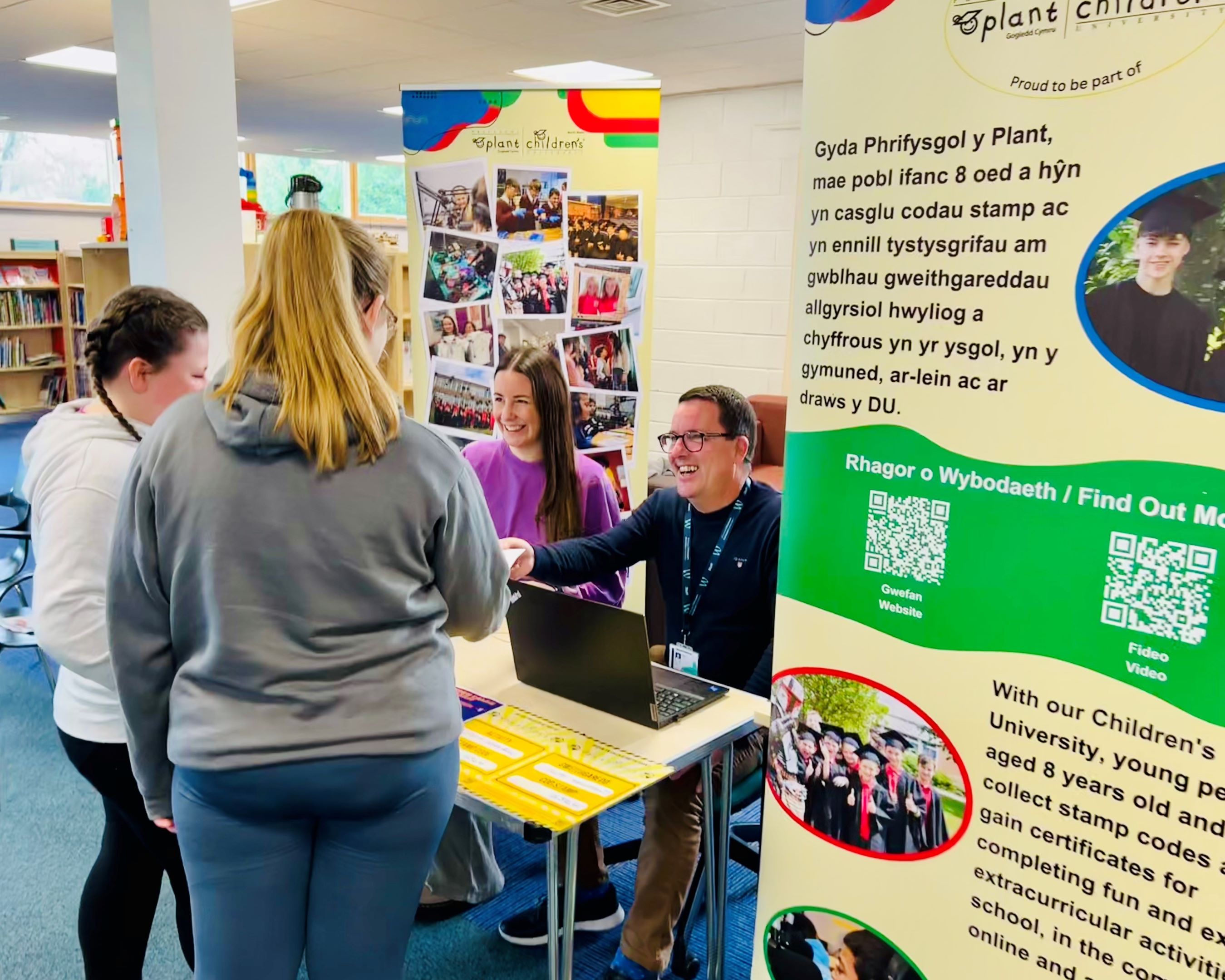 A photo of two people behind a desk with a children's university poster behind them giving information to two other people.