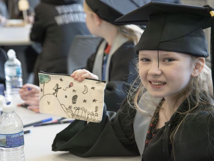 A young person in their children's university cap and gown showing off their pencil case they had drawn.