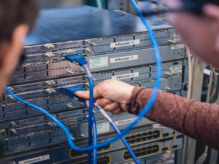 Two students face a server stack, one plugs in a cable