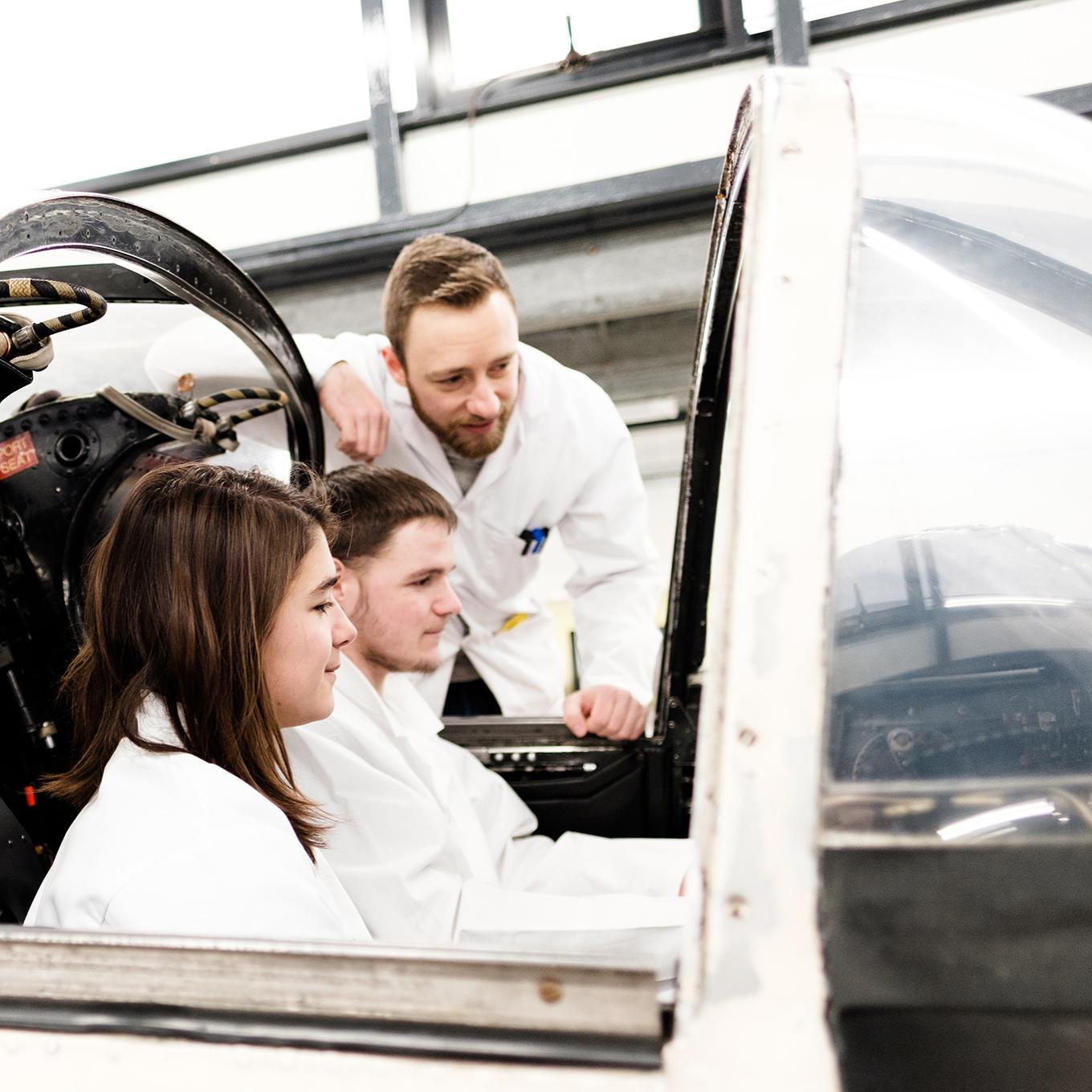 Two students sitting in the flight simulator, while another student leans in to look at the screens