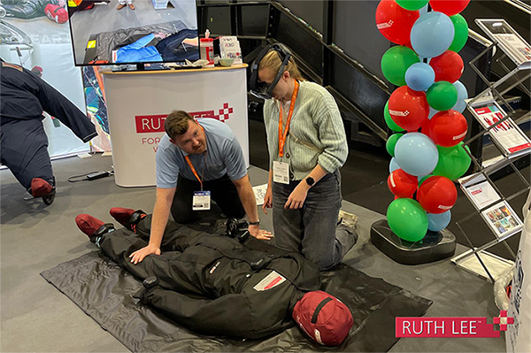 A mannequin lays on the ground as two people lean over it, one woman is wearing a virtual reality headset and a screen behind her shows the image she sees