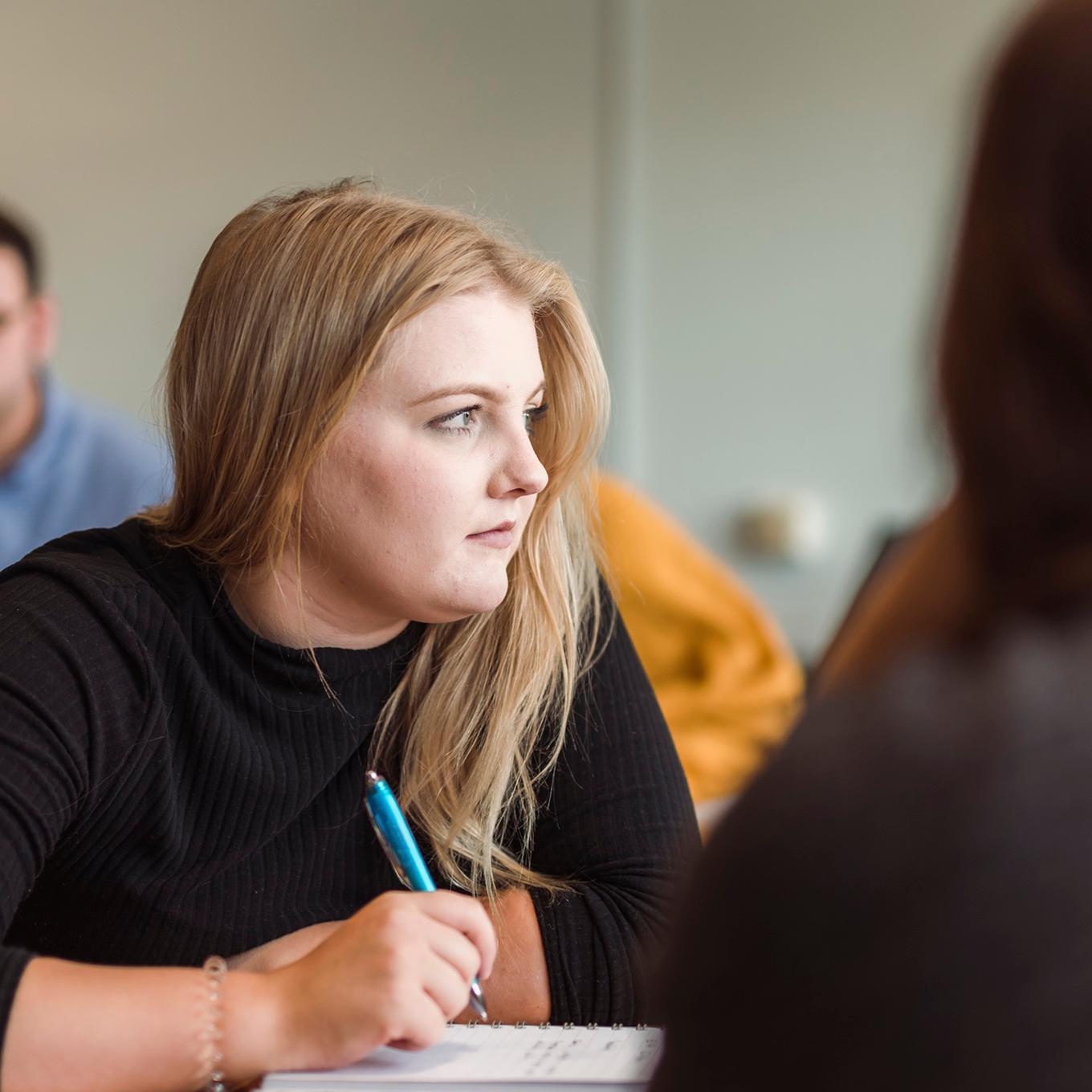 A student looks to the side, her pen poised over her notebook