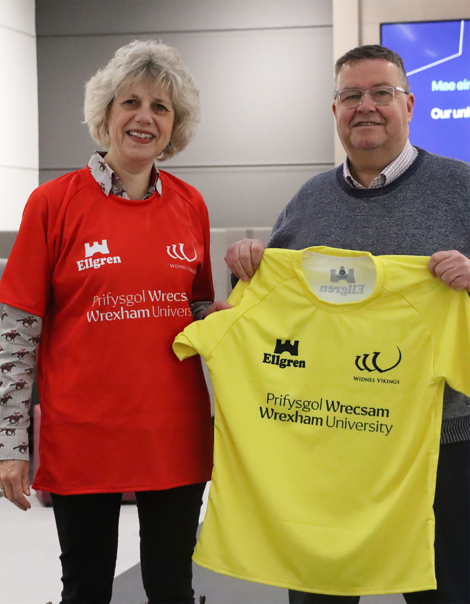 Professor Maria Hinfelaar wearing a red sports shirt and a yellow sports shirt held up next to her. Both tops have the Wrexham University logo on them