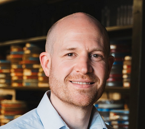 Head and shoulders shot of a male in a blue shirt with a blurred background of a library