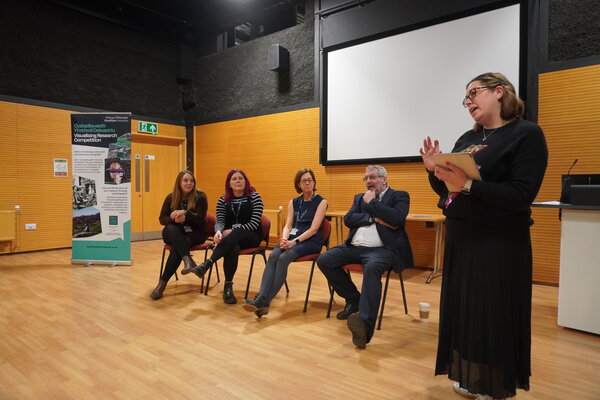 a panel of people sitting in a row with someone standing up chairing the panel