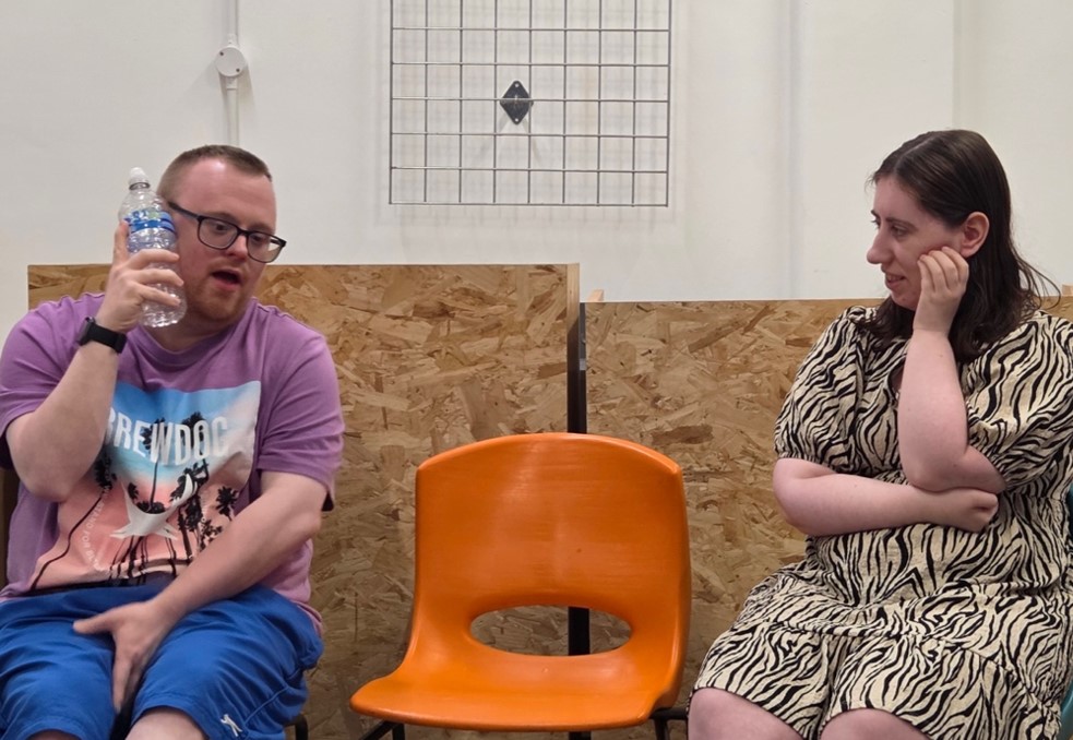 2 people sat on chairs, one holding a water bottle, with an empty seat between them