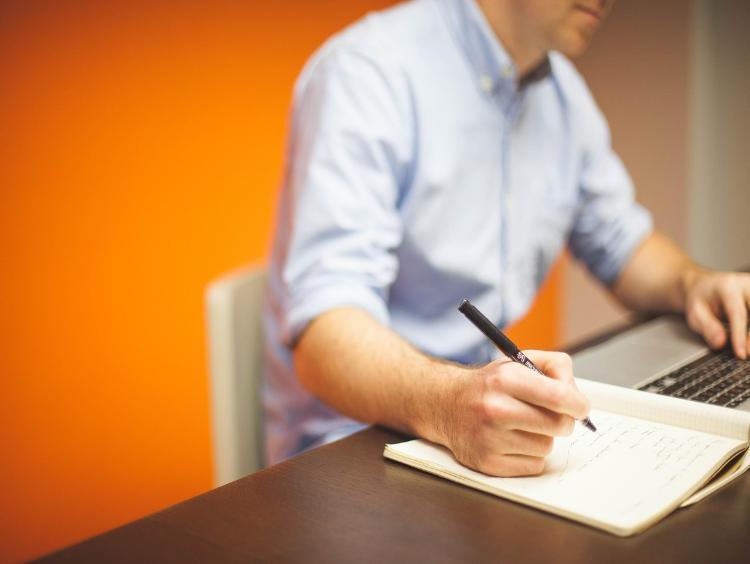 Orange wall in the background and person sat at a desk writing in the foreground